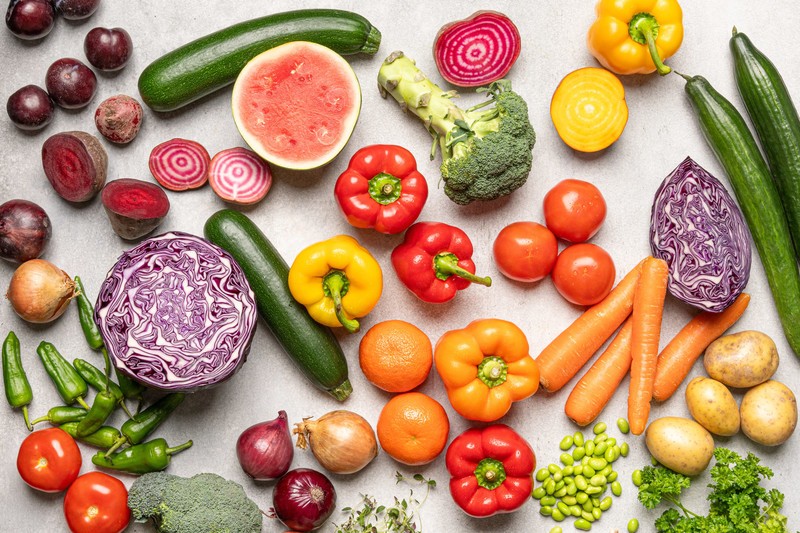 Värikkäitä vihanneksia ja hedelmiä, kuten paprikaa, porkkanoita, kurkkua, punakaalia, vesimelonia ja tomaatteja aseteltuna vaalealle pinnalle