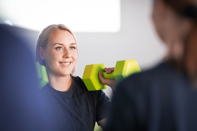 Fysioterapeutti nostaa vihreitä käsipainoja kuntosalilla, iloinen ilme kasvoillaan