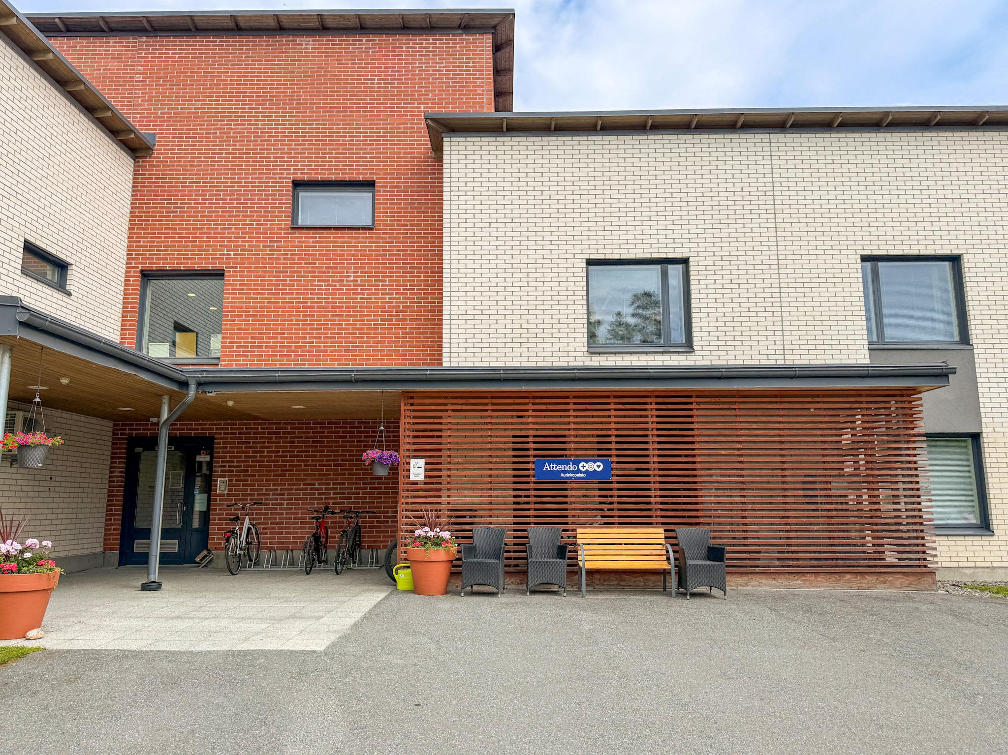 Kaksikerroksisen hoivakotirakennuksen julkisivu. Edustalla katos, istumapaikkoja ja kukkaruukku.