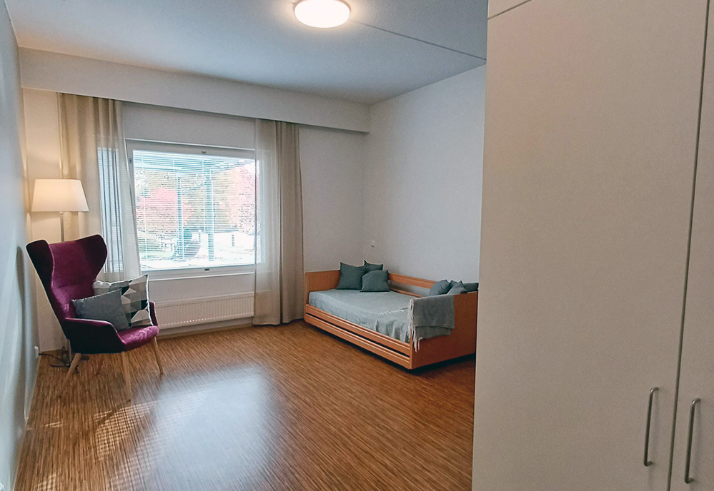 Asukashuone Riihivainiossa, puunvärinen lattia, nojatuoli ja sänky sekä ikkuna pihalle