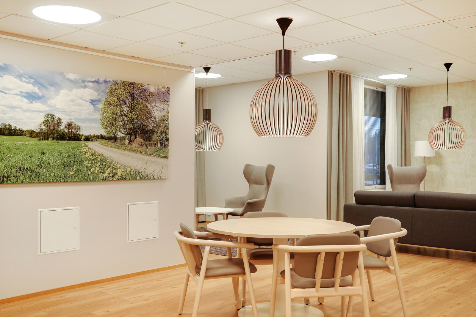 Moderni oleskelutila, jossa pyöreä ruokapöytä, vaaleat puiset tuolit, mukavat lepotuolit ja seinällä maisemataulu