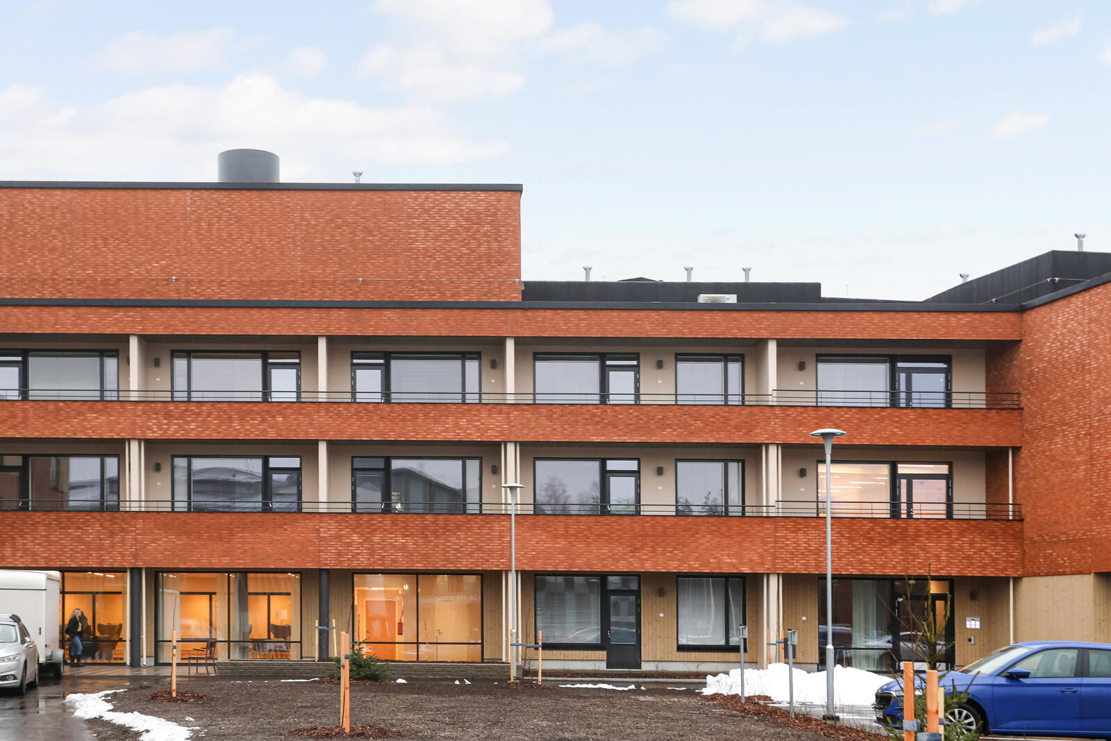 Salpakankaan punatiilinen kolmikerroksinen rakennus, jossa on parvekkeita jokaisessa kerroksessa. Etualalla luminen ja sora-alustainen piha