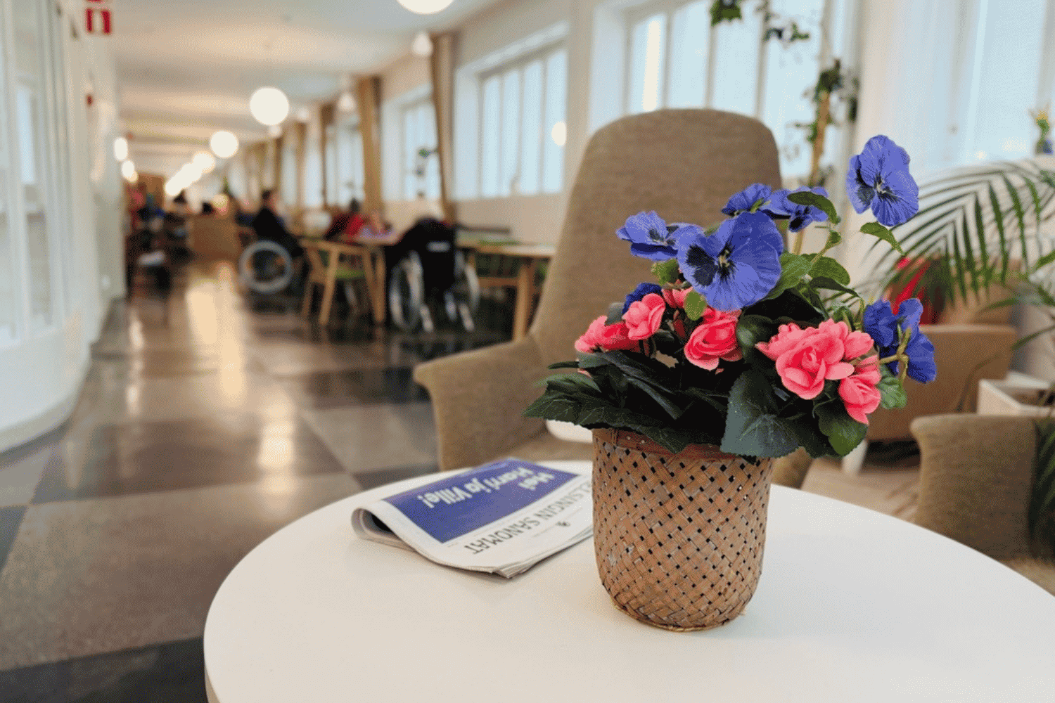 Attendo-hoivakodin valoisa yhteistila, jonka etualalla on pöydällä kukka-asetelma violeteilla ja vaaleanpunaisilla kukilla. Taustalla asukkaat istuvat yhdessä.