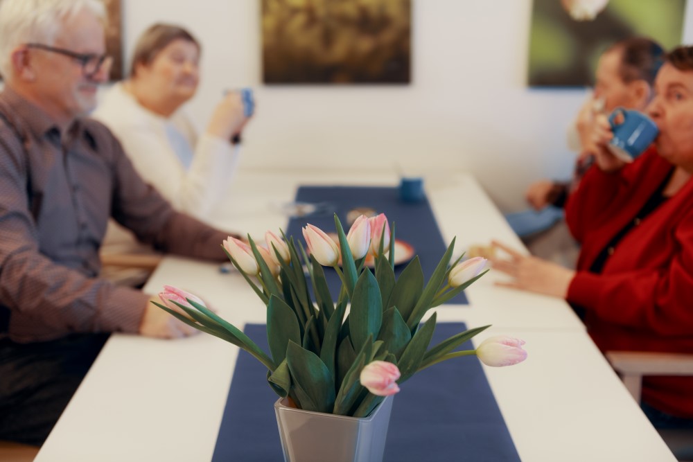 Vanhempia ihmisiä kahvilla pöydän ääressä, etualalla vaaleanpunaisia tulppaaneja maljakossa
