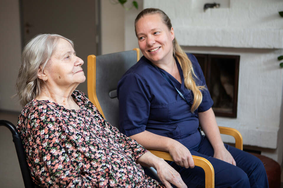harmaahiuksinen hoivakodin asukas ja pitkähiuksinen hoitaja istuvat vierekkäin juttelemassa