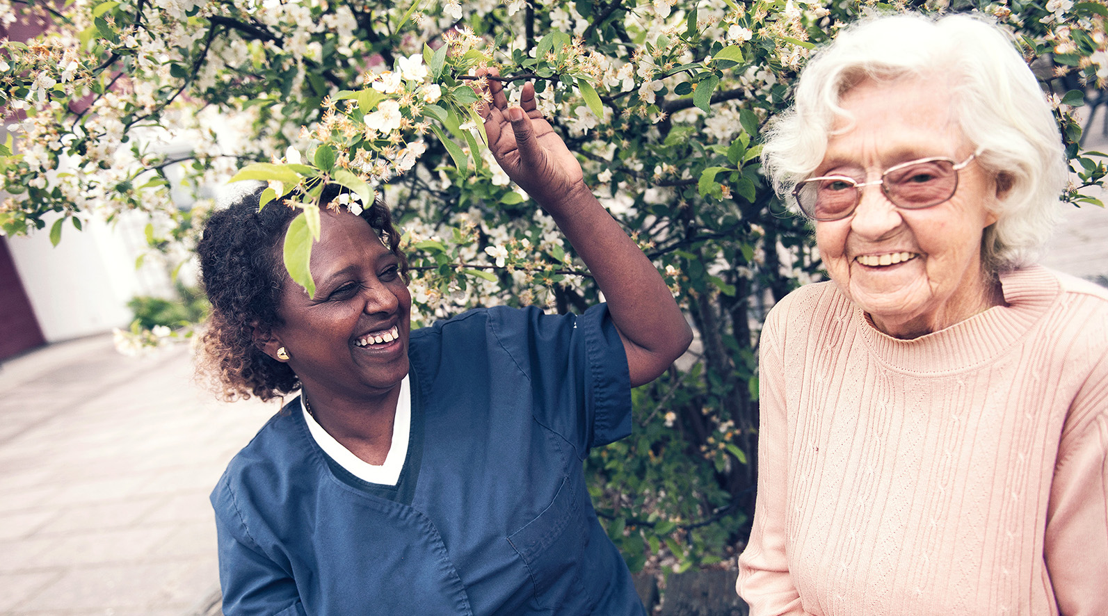 Hoitaja auttaa vanhusta ulkoilmassa kukkivien puiden vieressä