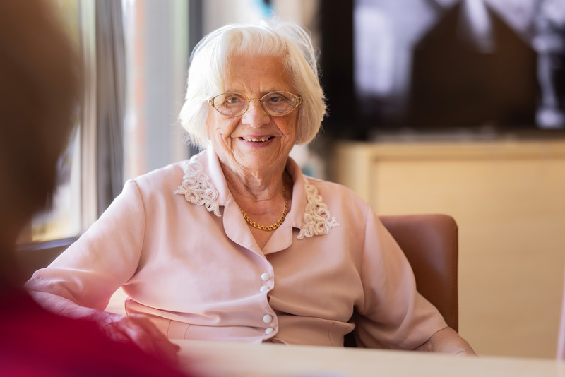 Harmaatukkainen nainen vaaleanpunaisessa paidassa istuu pöydän ääressä