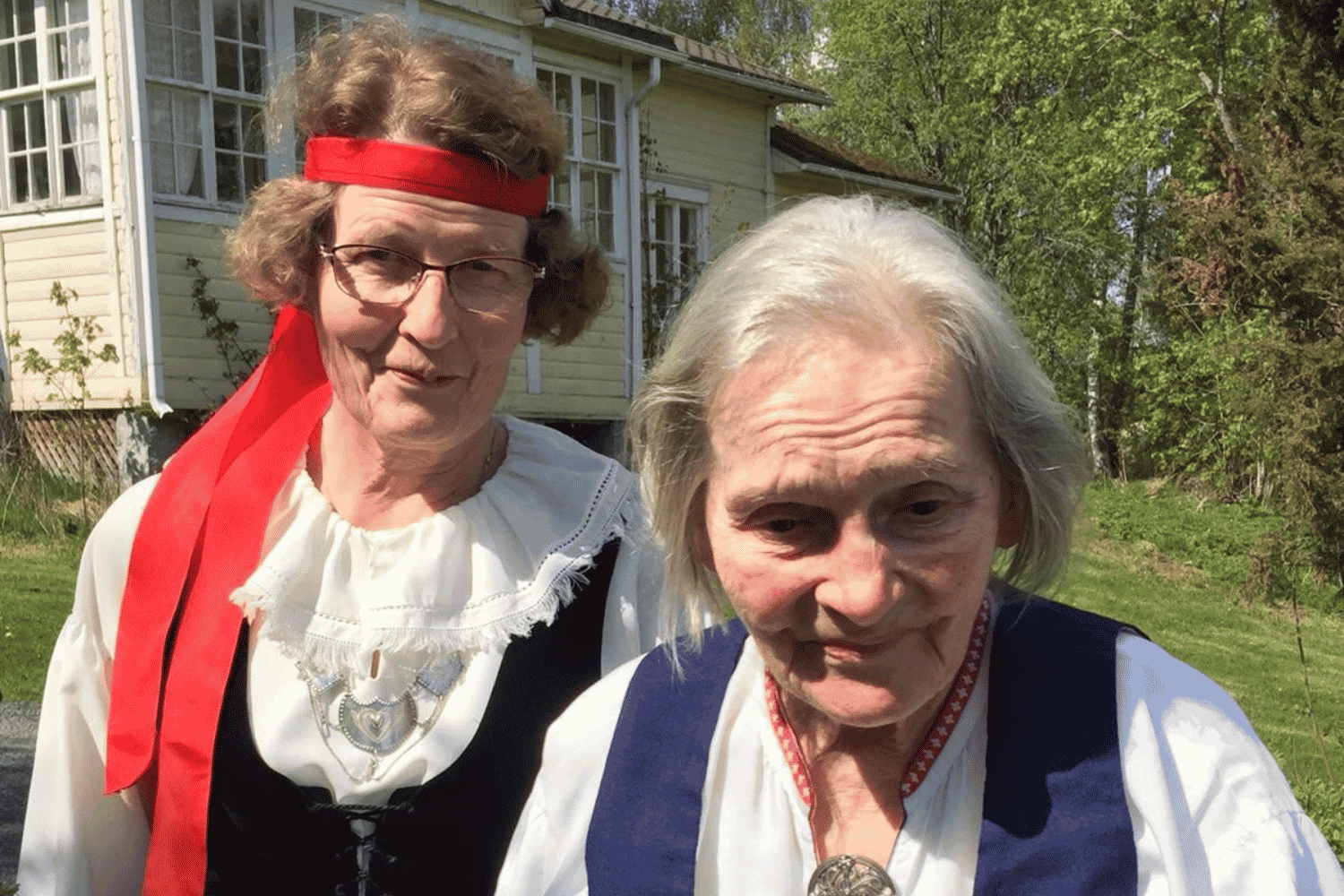 Läheinen ja asukas seisovat ulkona keltaisen puutalon edessä. Molemmilla on kansallispuvut. Läheisellä on punainen nauha hiuksissa.