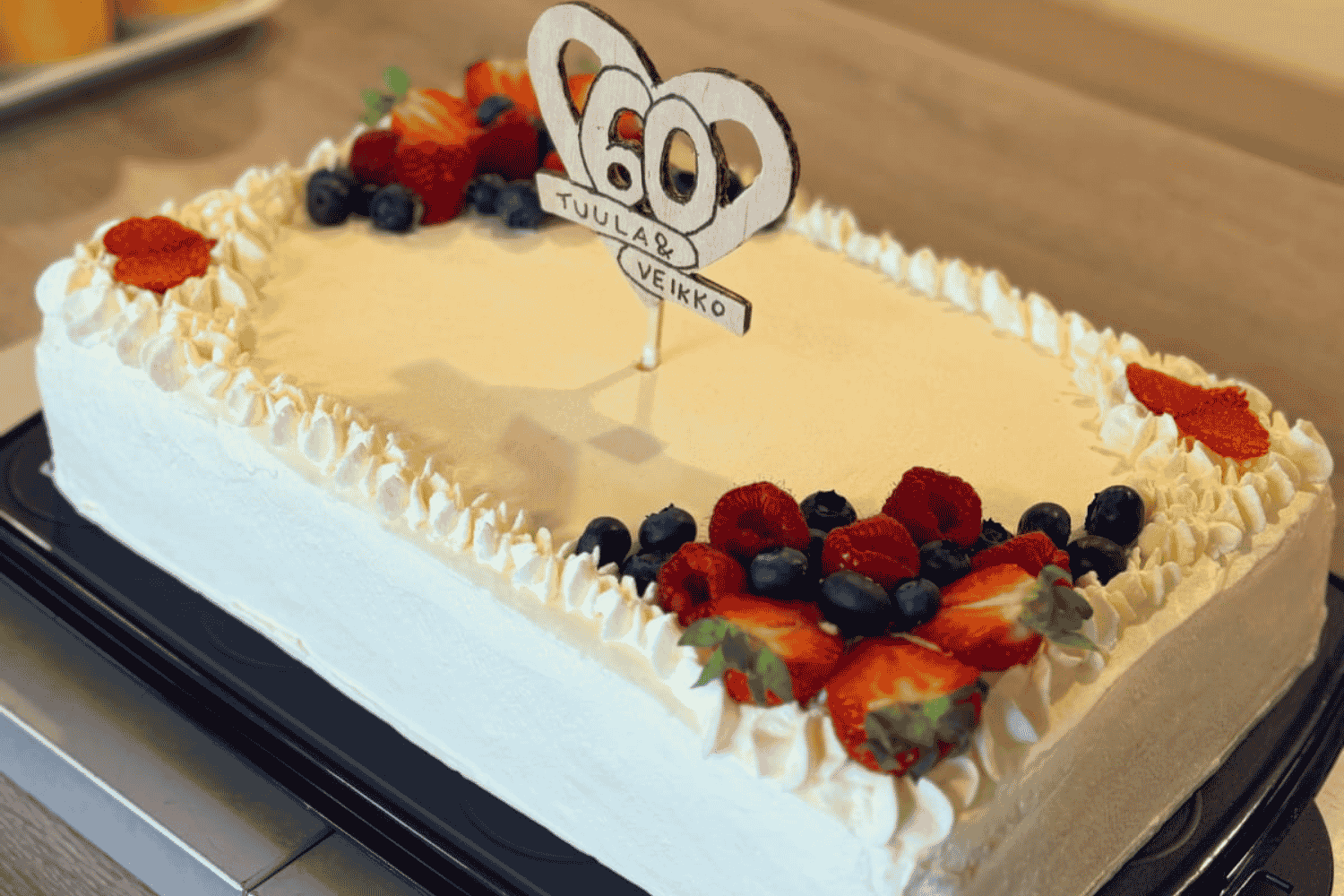 Juhlakakku, jonka päällä lukee "60, Tuula & Veikko". Kakun reunoilla on marjoja ja kermavaahtoa.