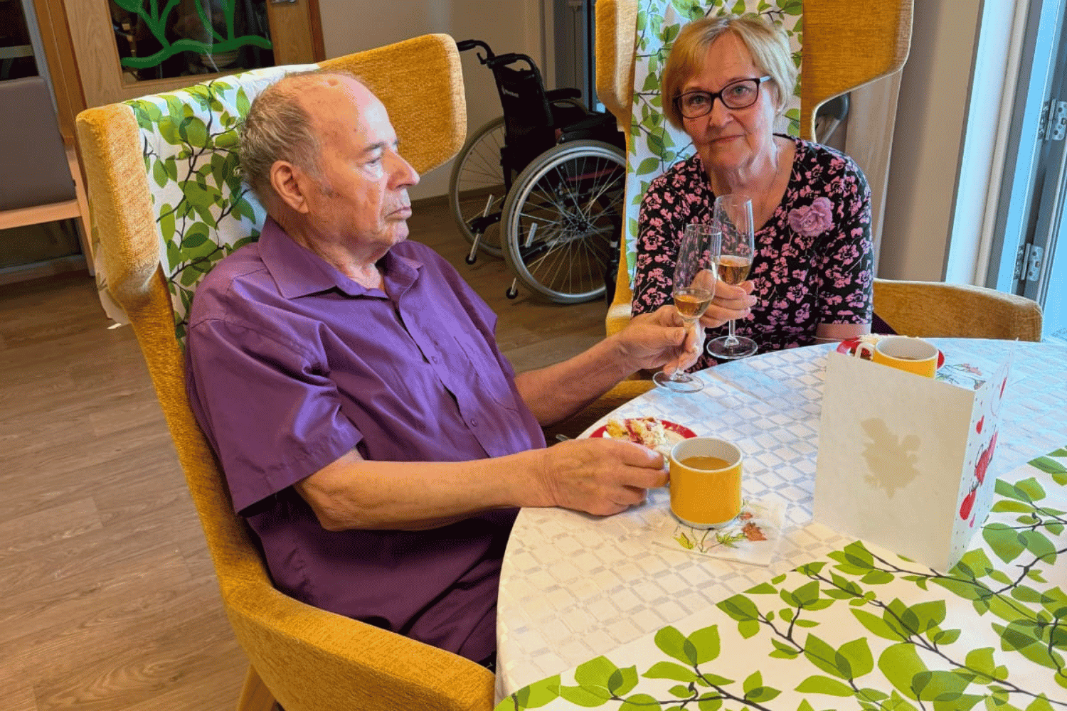 Veikko ja Tuula istuvat pöydässä ja kohottavat maljan. Pöydällä on kahvia ja kakkua.