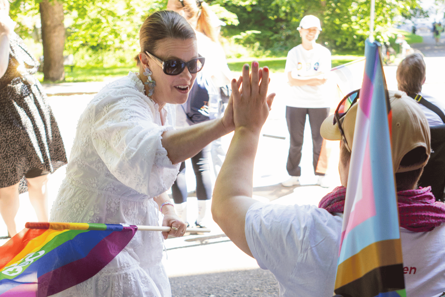 Kaksi ihmist&auml; ly&ouml; yl&auml;vitoset iloisesti Helsinki Pride -tapahtumassa. K&auml;siss&auml; sateenkaariv&auml;riset liput, taustalla hymyilevi&auml; ihmisi&auml; puiston valossa.