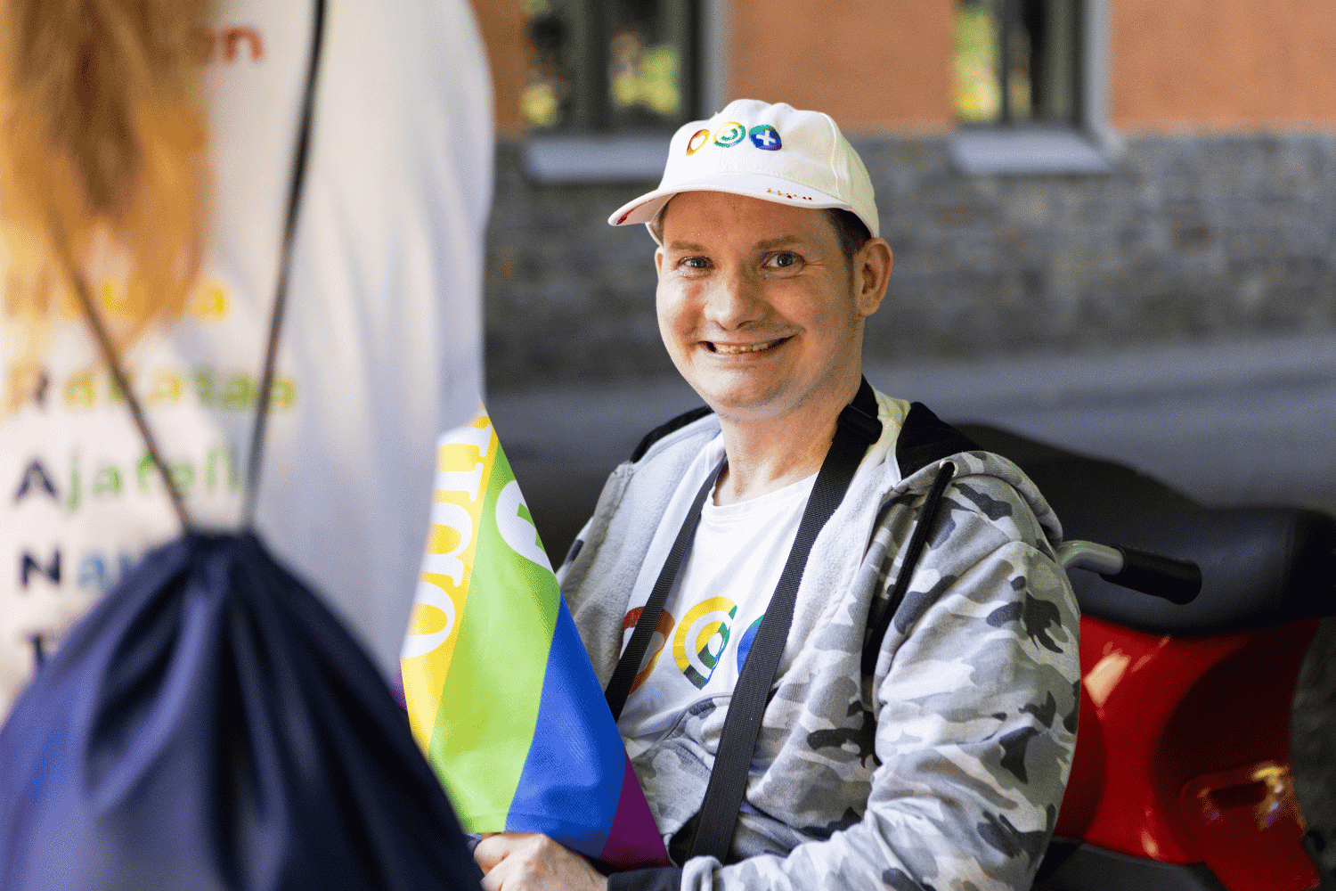Hymyilev&auml; asukas py&ouml;r&auml;tuolissa pit&auml;&auml; sateenkaariv&auml;rist&auml; lippua. P&auml;&auml;ss&auml; valkoinen Attendo-lippalakki. 