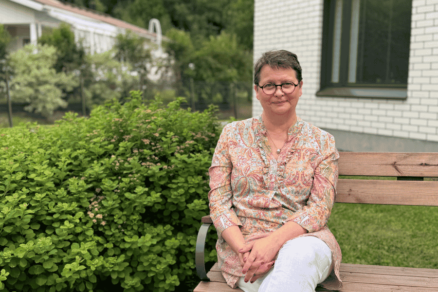 Merja istuu hoivakodin pihalla ulkona ja hymyilee kameraan.