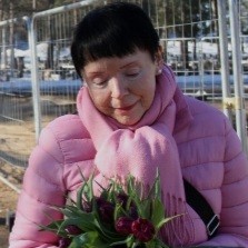Muistisairaan l&auml;heinen Tarja seisoo hymyillen vaaleanpunaisessa takissa tulppaanit k&auml;dess&auml;&auml;n.