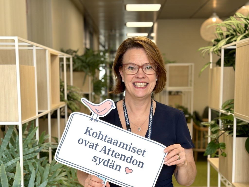Virpi hymyilee kameralle ja pitää kylttiä, joissa lukee "Kohtaamiset ovat Attendon sydän".