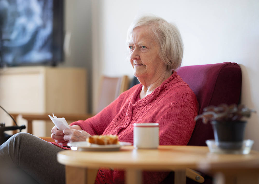 Iäkäs nainen istuu nojatuolissa, jolla on punainen neule. Hän pitää paperia kädessään. Hänen vieressään on pöytä, jossa on kahvikuppi ja pullaa.