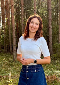 Iida katsoo suoraan kameraan kesäisessä mäntymetsässä