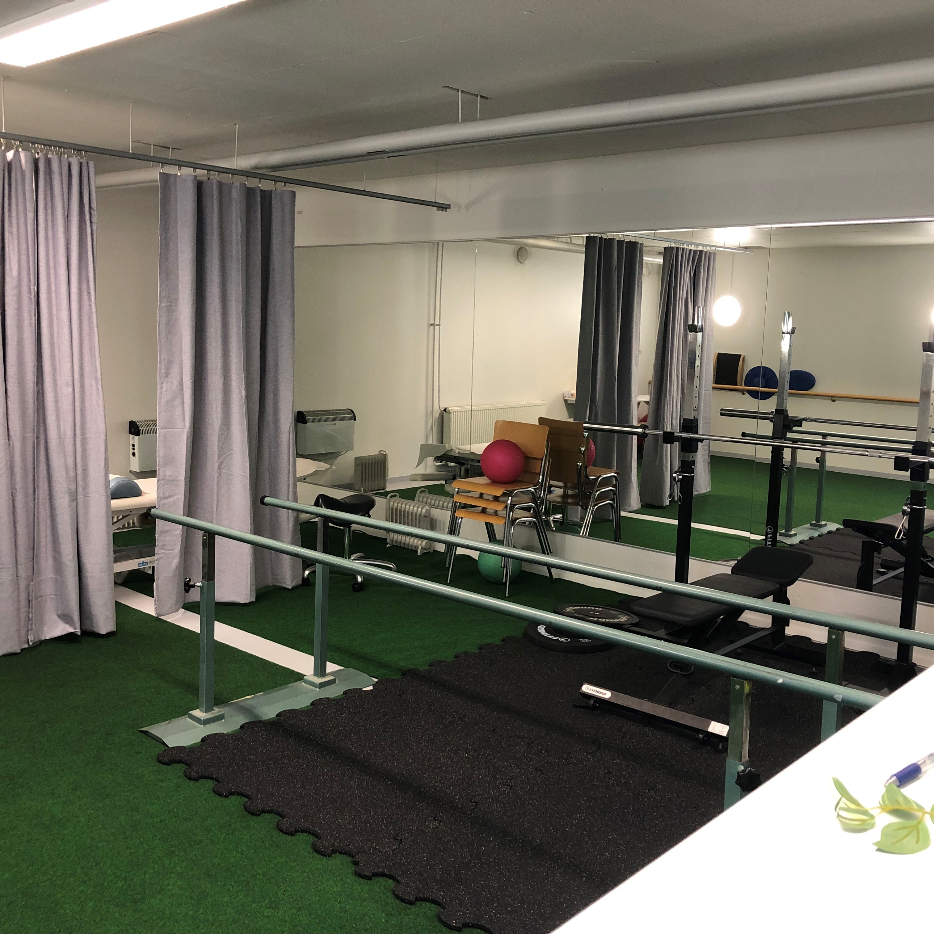 Kuntosali, jossa on peili, kuntosalilaitteita, tangot ja kuntopallo, vihreä lattiamatto ja harmaat verhot