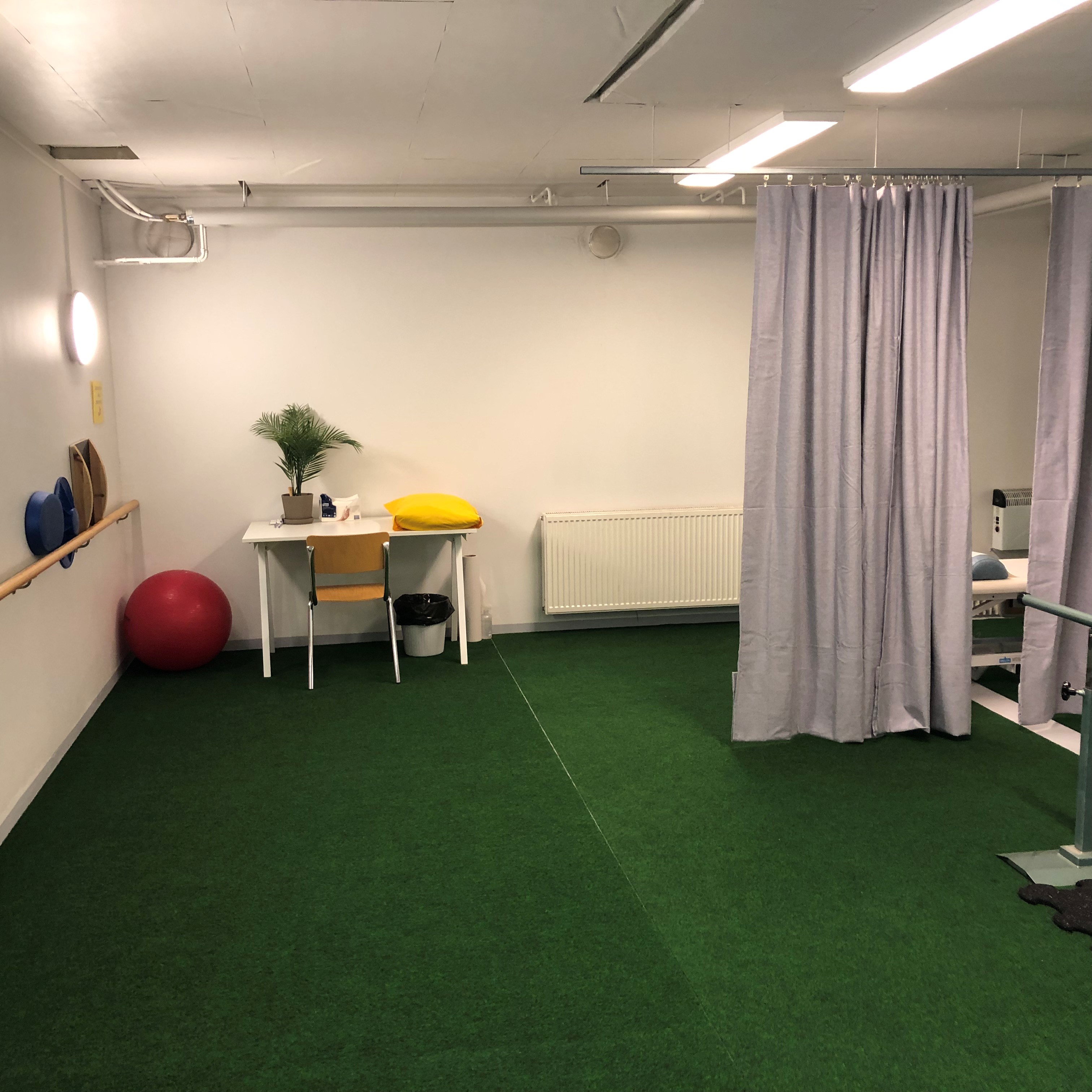 Fysioterapiatila vihreällä matolla, pöytä kasvin ja keltaisen tyynyn kanssa, punainen jumppapallo ja harmaat verhot
