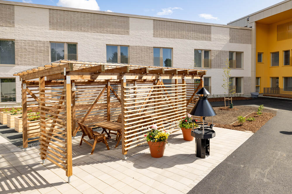 Puutarhakatoksen alla pöytä ja tuolit, vieressä grilli, nykyaikaisella pihalla kerrostalon edustalla