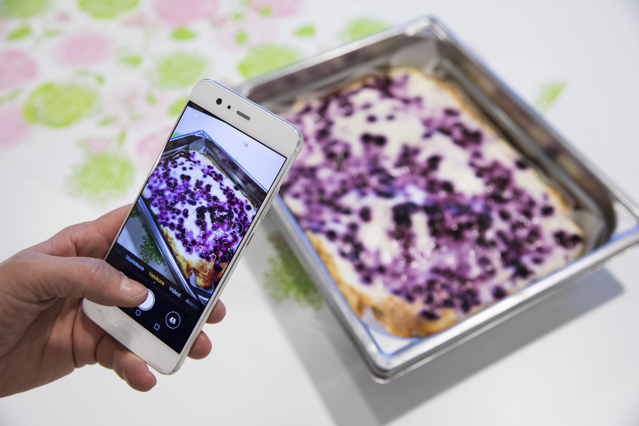 Pöydällä mustikkapiirakka suorakaiteen muotoisessa vuoassa, ja siitä otetaan kännykällä kuvaa